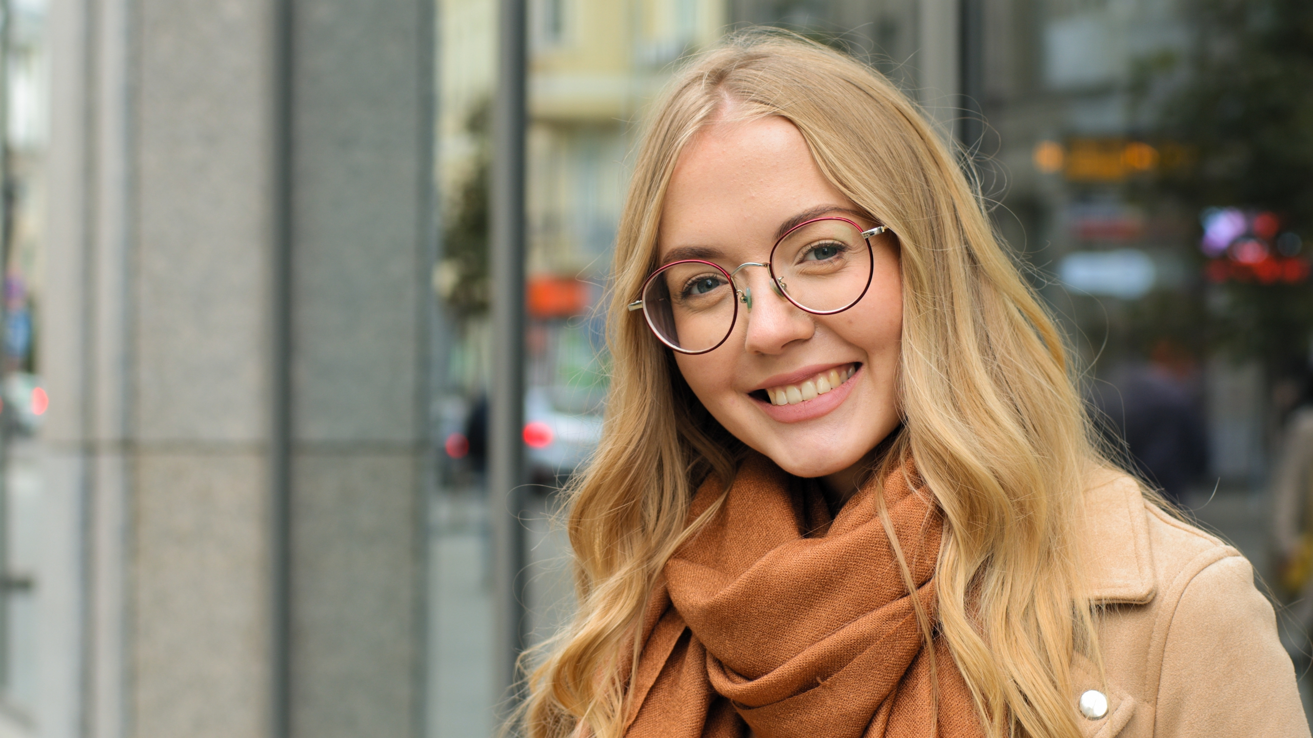 Happy woman wearing round eyeglasses and a scarf while standing outdoors in an urban setting, symbolizing clear vision, comfort, and fashionable eyewear options – glasses in plymouth