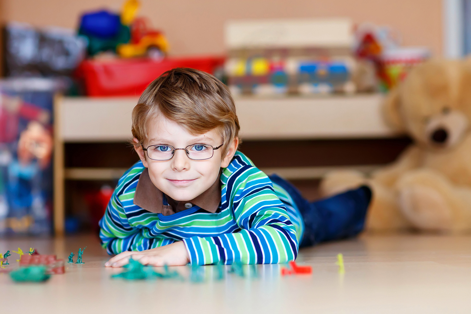 A young boy wearing glasses and a striped shirt lies on the floor, smiling as he plays with colorful toy soldiers in a playroom filled with stuffed animals and educational toys. - Kennedy Vision Health Center