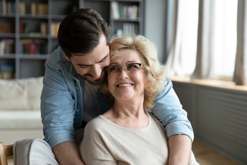 Smiling senior woman embraced by a loved one representing trusted, compassionate vision care – Eye Doctor Wayzata MN Happy older woman wearing glasses smiling while embraced by a family member at home, symbolizing comfort, trust, and family-centered eye care – Eye Doctor Wayzata MN