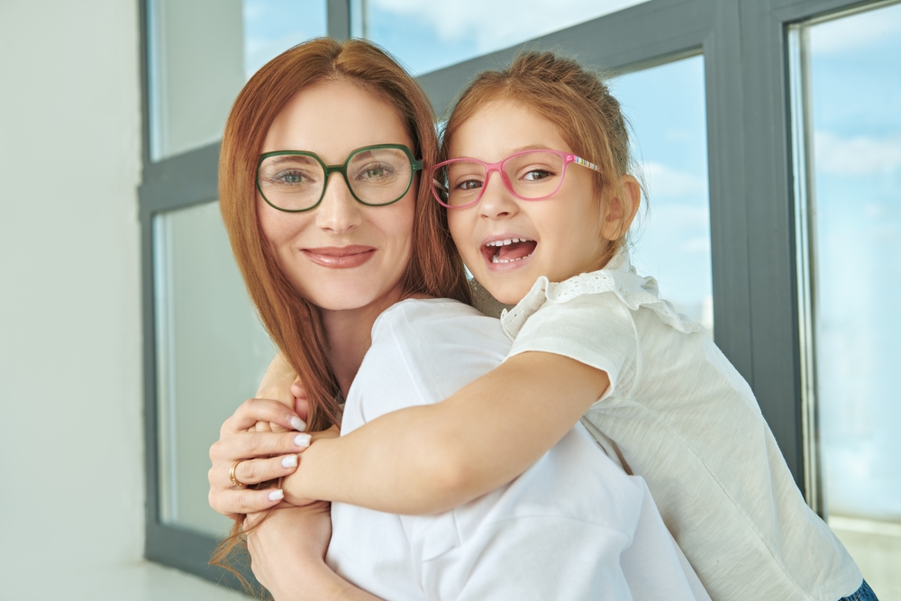 Mother and daughter wearing glasses smiling together representing family-focused vision care – Eye Doctor Wayzata MN Smiling mother and young daughter wearing eyeglasses and embracing indoors, symbolizing trusted, compassionate eye care for families – Eye Doctor Wayzata MN
