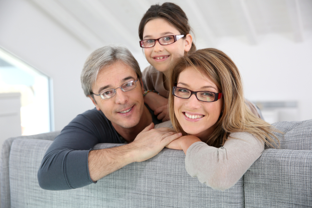 Happy family of three wearing eyeglasses and smiling on a couch, symbolizing trusted, compassionate vision care for patients of all ages – Eye Doctor Plymouth