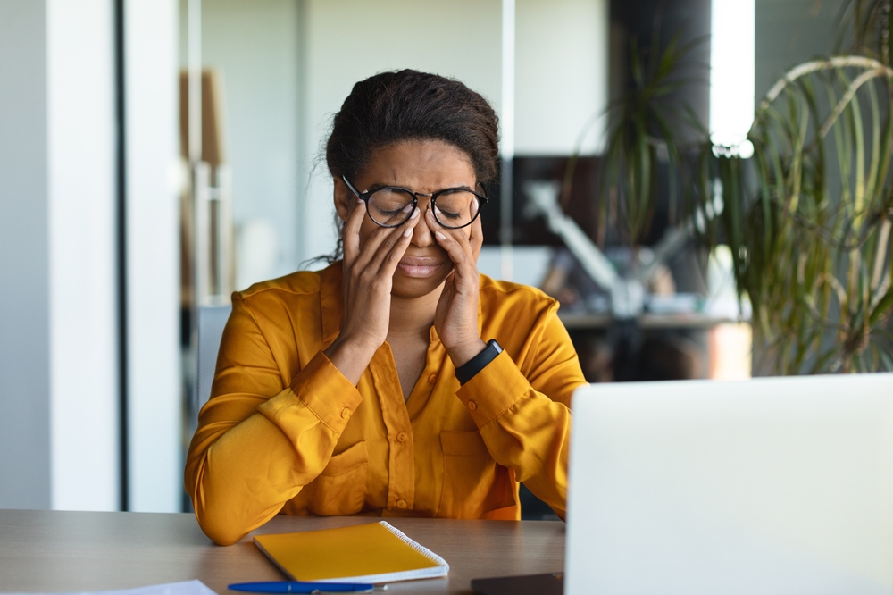 Woman rubbing her eyes while seated at a desk with a laptop, representing digital eye strain symptoms and the importance of comprehensive eye exams – eye exam plymouth