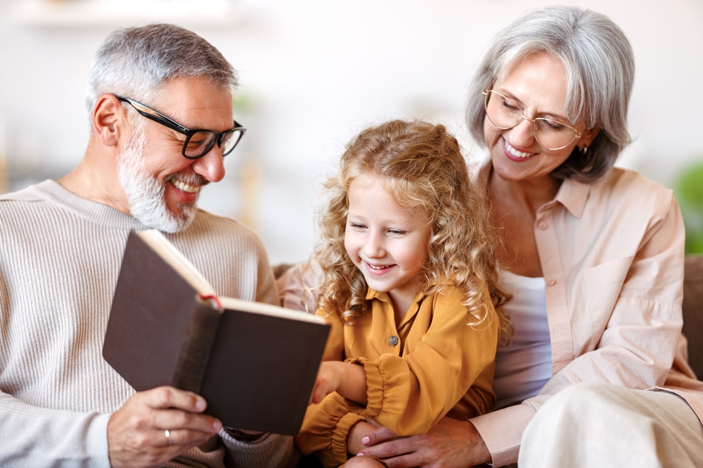 A smiling grandfather with glasses reads a book to his curly-haired granddaughter while the grandmother, also wearing glasses, watches warmly beside them. - Kennedy Vision Health Center